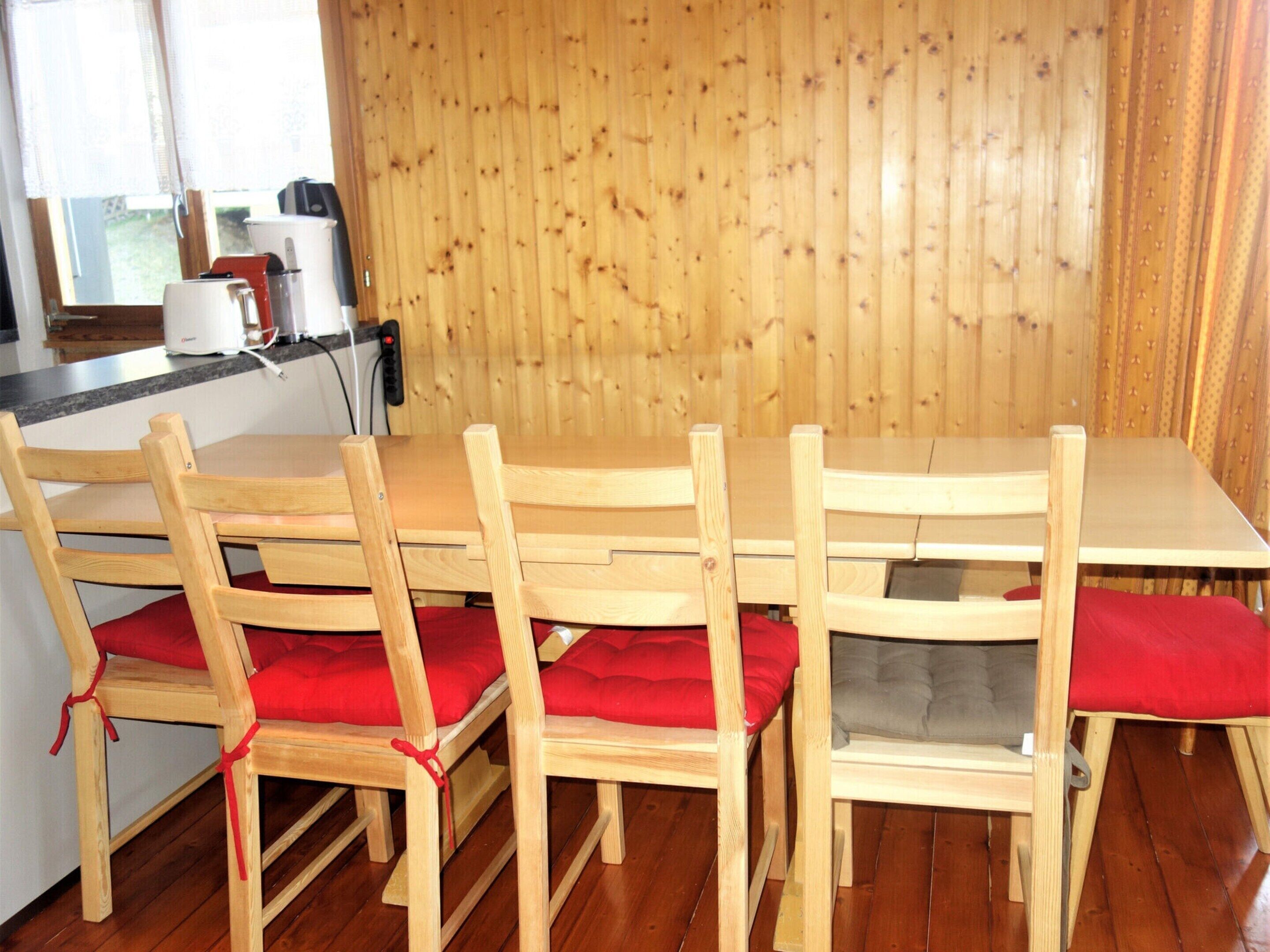 Una mesa de comedor de madera con cuatro sillas, cada una con un cojín rojo. La habitación tiene paneles de madera en las paredes y una ventana con cortina.