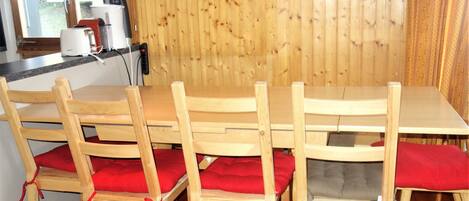 Una mesa de comedor de madera con cuatro sillas, cada una con un cojín rojo. La habitación tiene paneles de madera en las paredes y una ventana con cortina.