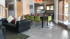 Een moderne woonkamer met een zwarte leren bank, groene fauteuils en een eettafel voor zes personen. Achtergrond toont een keuken met grijze kastjes en een houten plafond.
