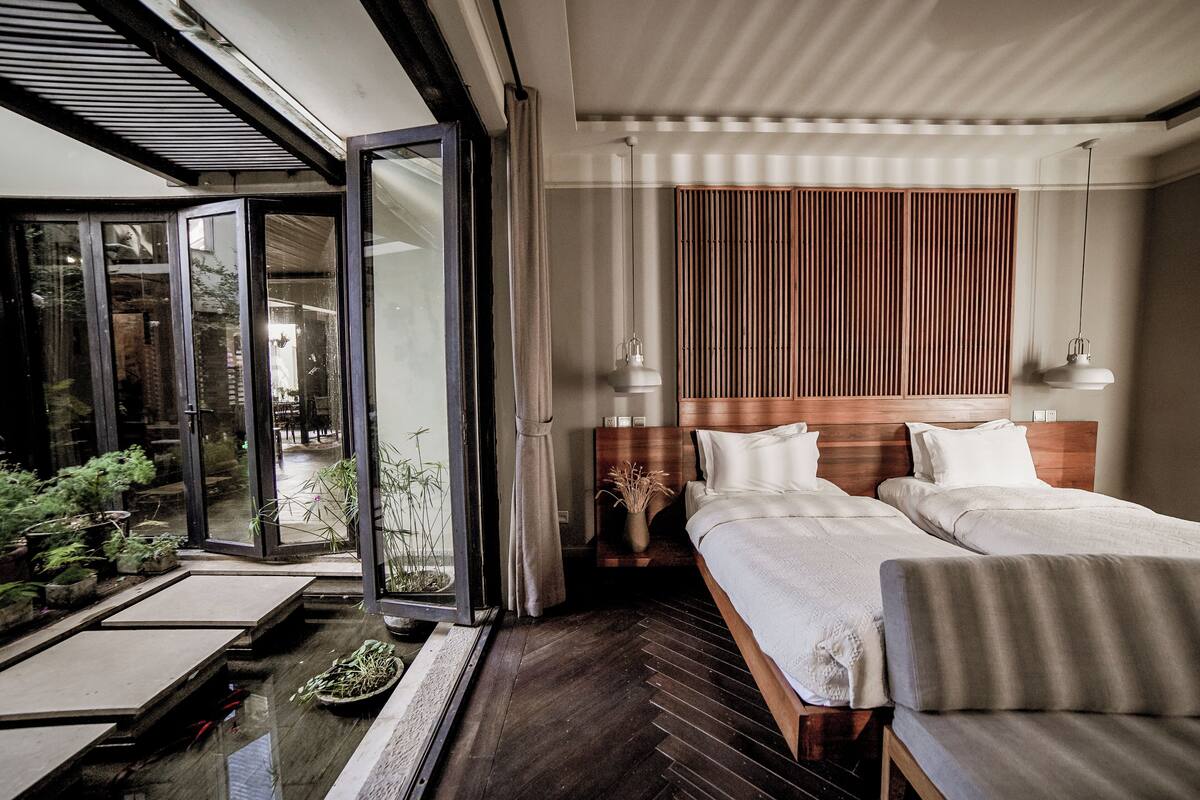 A modern hotel room with a large sliding glass door leading to a balcony, a wooden headboard, two beds, a small table with a vase, and a sofa.