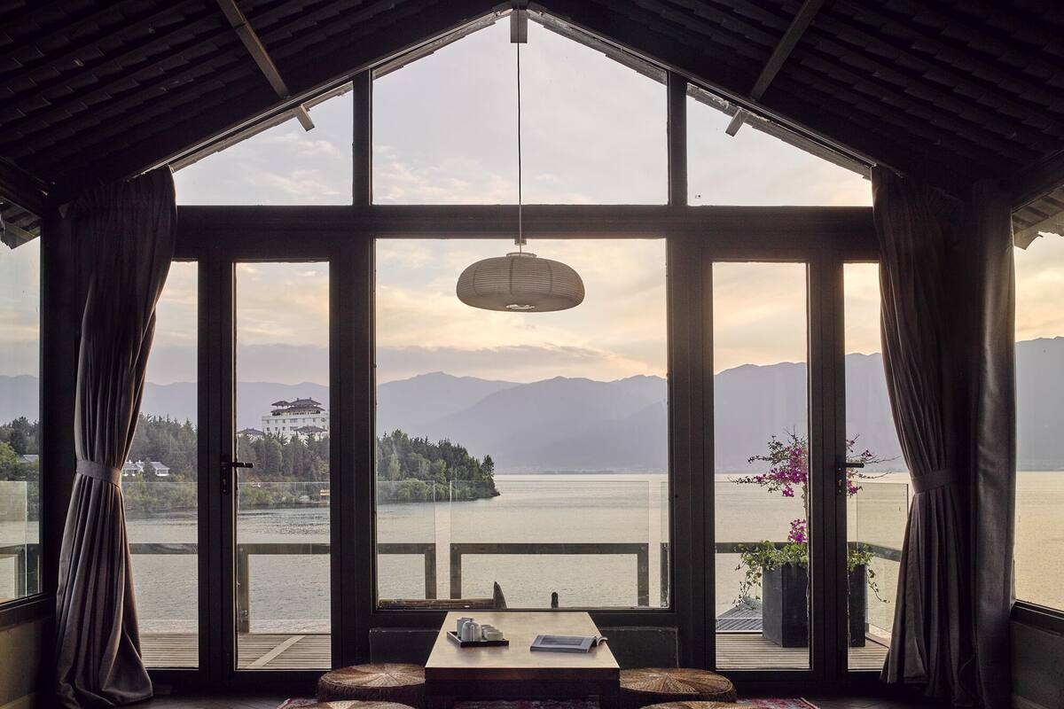 A room with a large window overlooking a lake and mountains, wooden furniture, and a hanging light fixture.