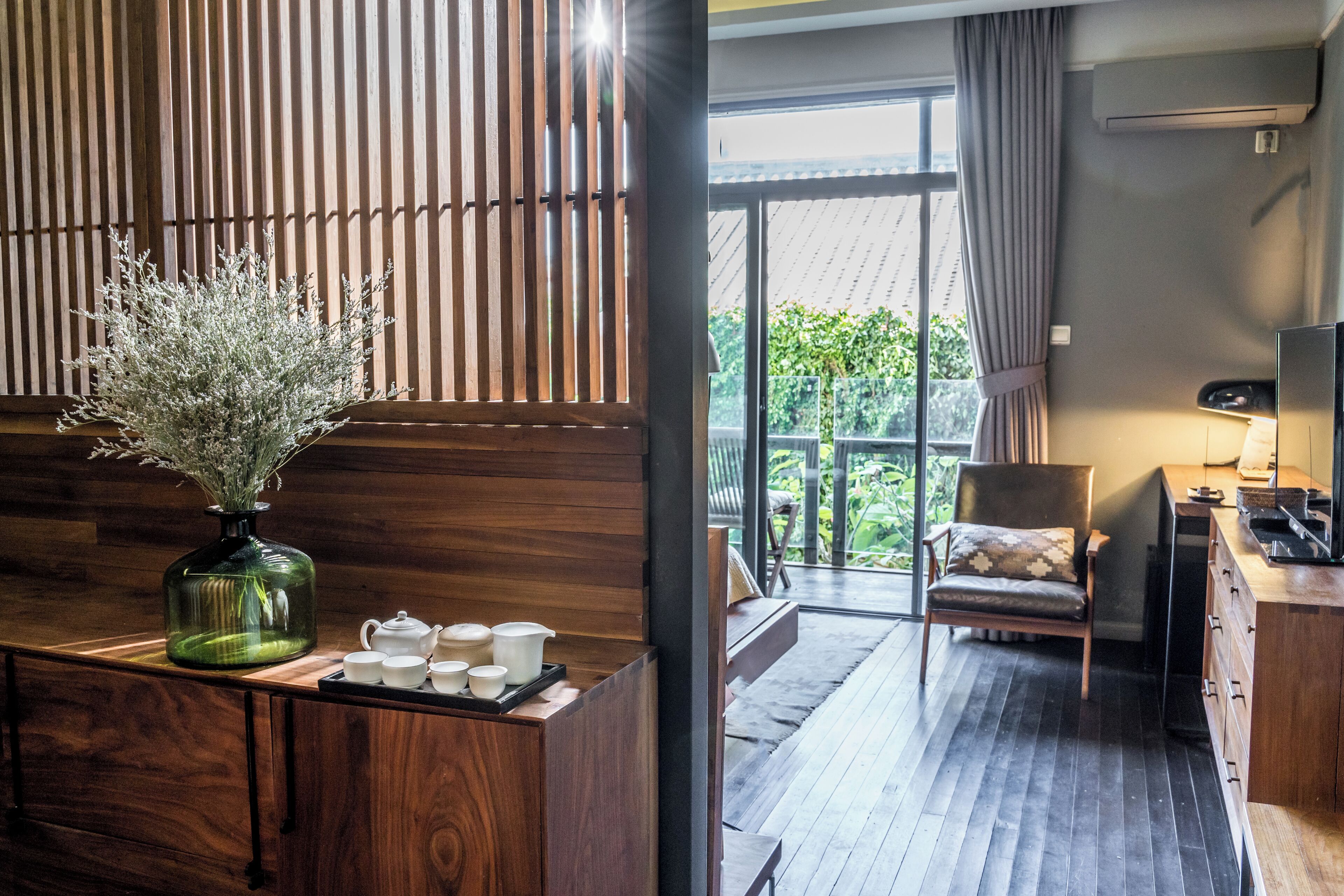 Une chambre d’hôtel moderne avec un comptoir d’accueil en bois, un vase contenant des fleurs séchées, un service à thé et une vue sur l’extérieur grâce à une porte coulissante.