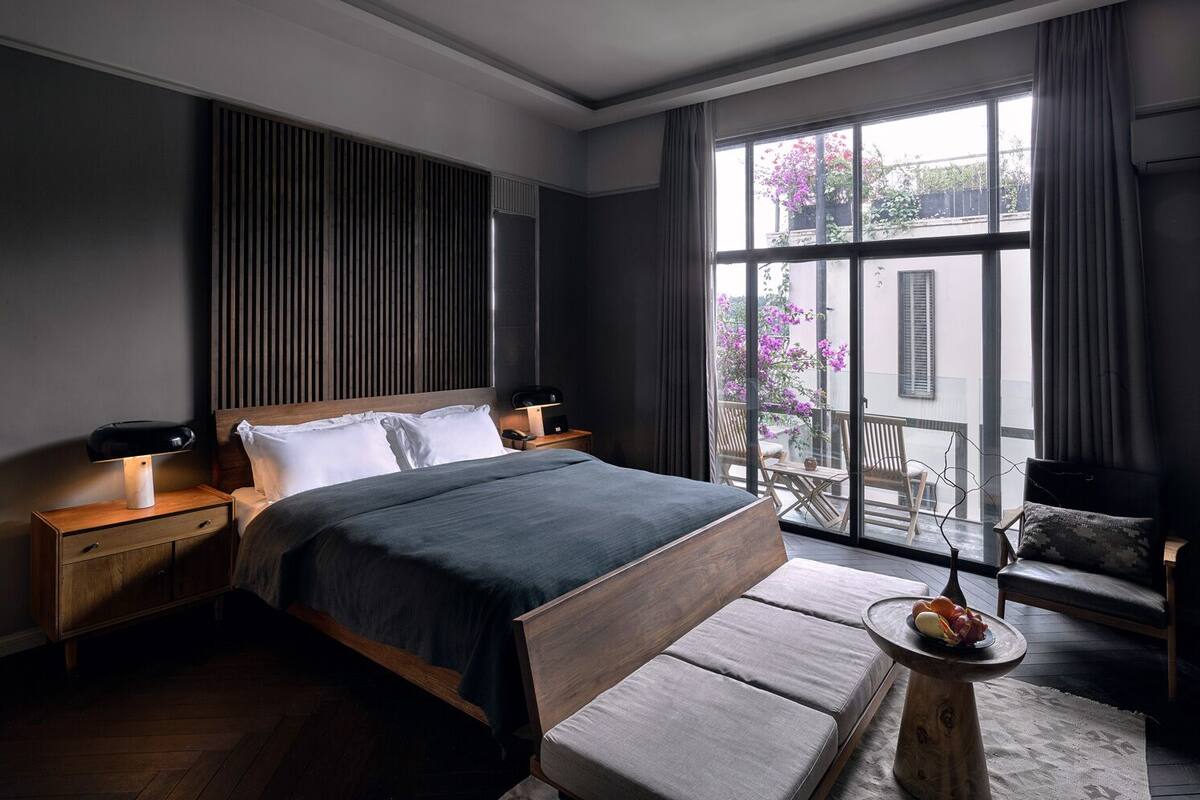 A modern bedroom with a large bed, wooden headboard, bedside tables, and a view of the outside balcony.