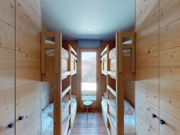 Une cabane en bois avec des lits superposés disposés le long d’un couloir central, une petite table ronde et une vue sur un paysage montagneux par la porte.