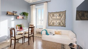 A living room with a white sofa, a dining table with chairs, a chandelier, and a large tapestry on the wall.