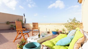 A rooftop terrace with a wooden chair, a cushioned sofa, potted plants, and a clear blue sky.