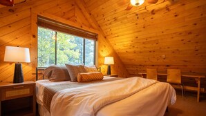 A cozy bedroom with a large bed, two bedside tables with lamps, a window with a view of trees, and a wooden ceiling. - Family-Friendly Modern Cabin with Spectacular Views (Angel Fire)