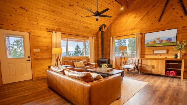 A cozy cabin interior with a leather sofa, a fireplace, and a ceiling fan. - Family-Friendly Modern Cabin with Spectacular Views (Angel Fire)