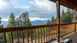 A wooden deck with a glass table and chairs, overlooking a mountainous landscape with trees. - Family-Friendly Modern Cabin with Spectacular Views (Angel Fire)