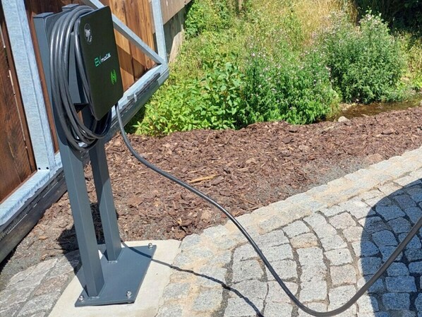 An electric vehicle charging station with a cable connected to a vehicle, set against a backdrop of a garden with mulch and greenery. - Spacious Villa With Good Ambience (Cerny Dul)