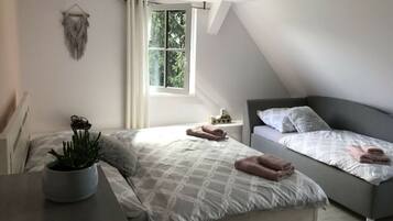 A bedroom with a bed, a bedside table with a plant, a window with curtains, and a small bedside table with a lamp.