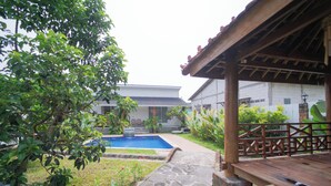 A pool area with stone pathways, a wooden deck, and a building with a tiled roof in the background. - Villa Sawah Prambanan (Prambanan)