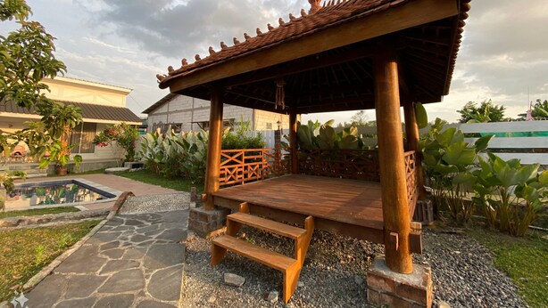 A wooden pavilion with a tiled roof, surrounded by gravel and plants, with a pool and a building in the background. - Villa Sawah Prambanan (Prambanan)