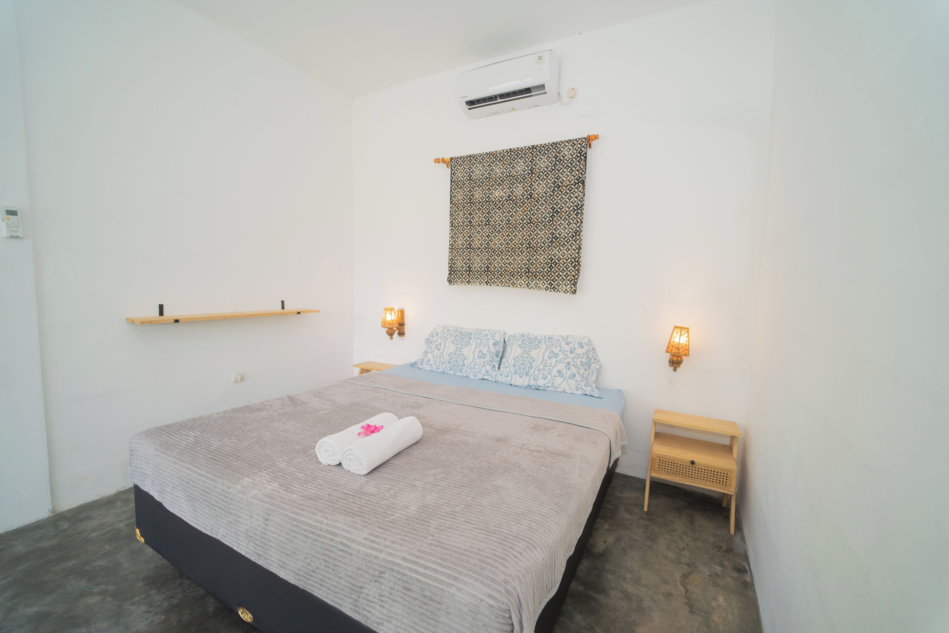 Un dormitorio con una cama, aire acondicionado de pared, un tapiz estampado, dos lámparas de noche y un pequeño estante de madera.