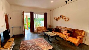 A living room with a sofa, coffee table, and TV. There is a sliding glass door leading to a balcony with a view of greenery. - Villa Sawah Prambanan (Prambanan)