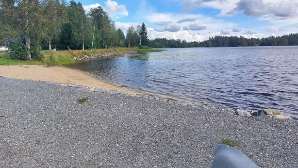 Une plage de galets bordant un lac paisible, entourée d’arbres et offrant une vue sur un ciel dégagé.