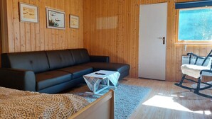 Une pièce chaleureuse avec un décor intérieur en bois, un canapé noir, une petite table recouverte d’une couverture, un fauteuil à bascule et une porte donnant sur l’extérieur.