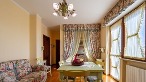 Una sala de estar acogedora con un sofá estampado, una mesa de comedor de madera con sillas, una araña y cortinas con motivos florales.