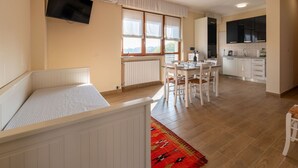 Ein kleines Zimmer mit Bett, Esstisch mit Stühlen, Küchenzeile und einem an der Wand montierten Fernseher.