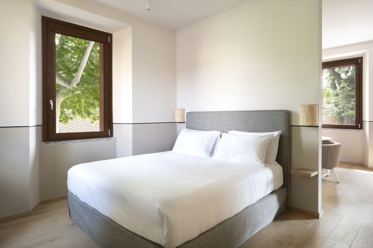 A modern bedroom with a large bed, a window with a view of trees, and a bedside table with a lamp.
