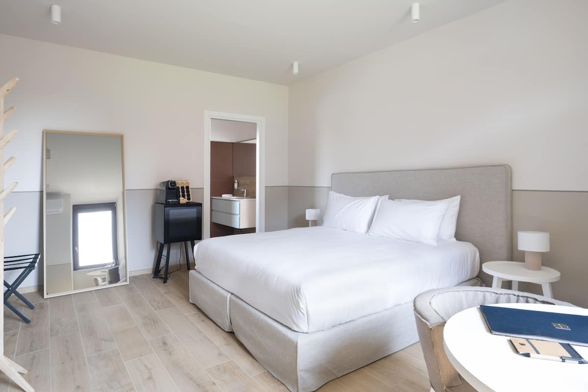 A modern hotel room with a large bed, bedside tables, a desk with a chair, and a mirror.