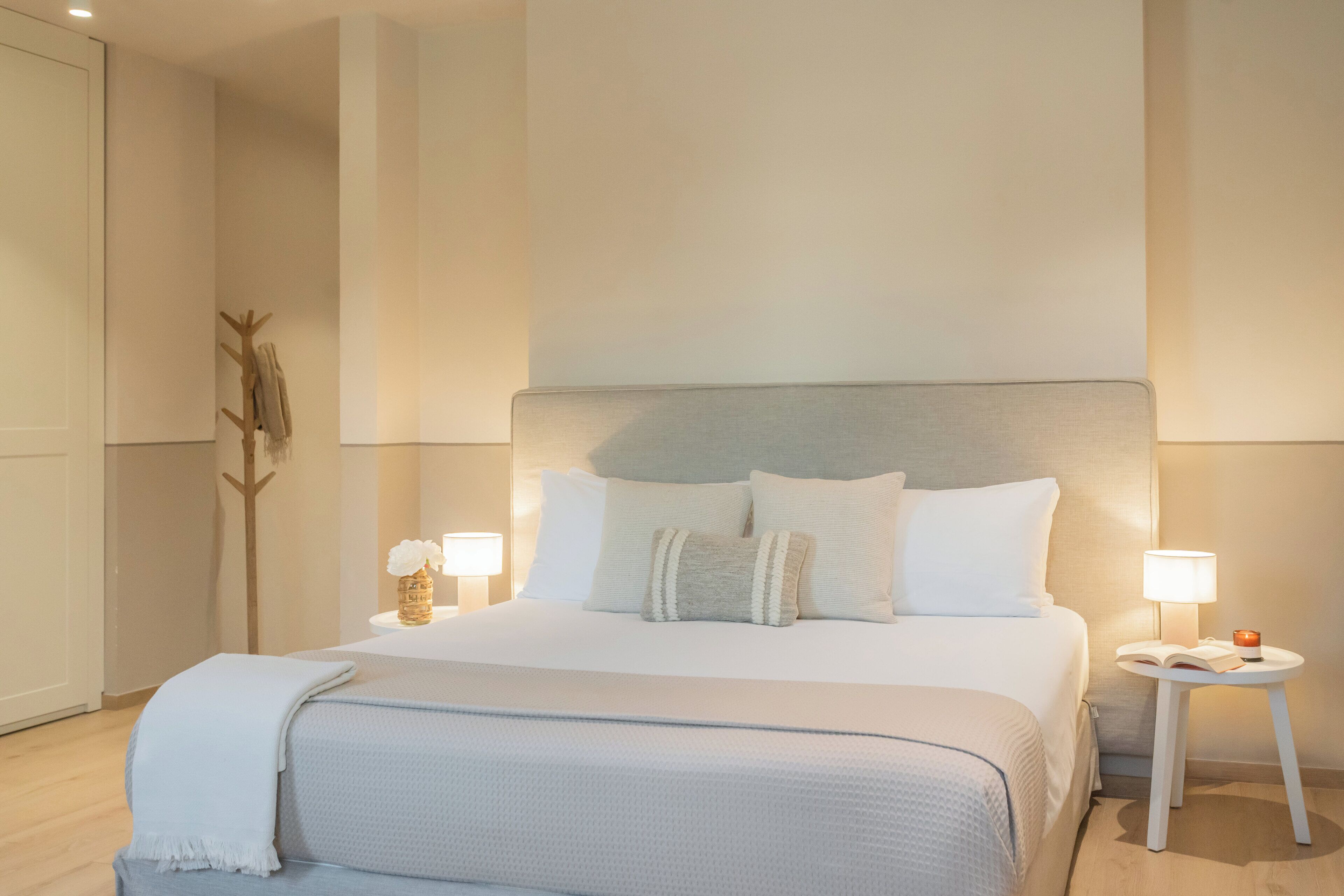 A modern hotel room with a large bed, two bedside tables with lamps, a small table with a vase, and a coat rack.