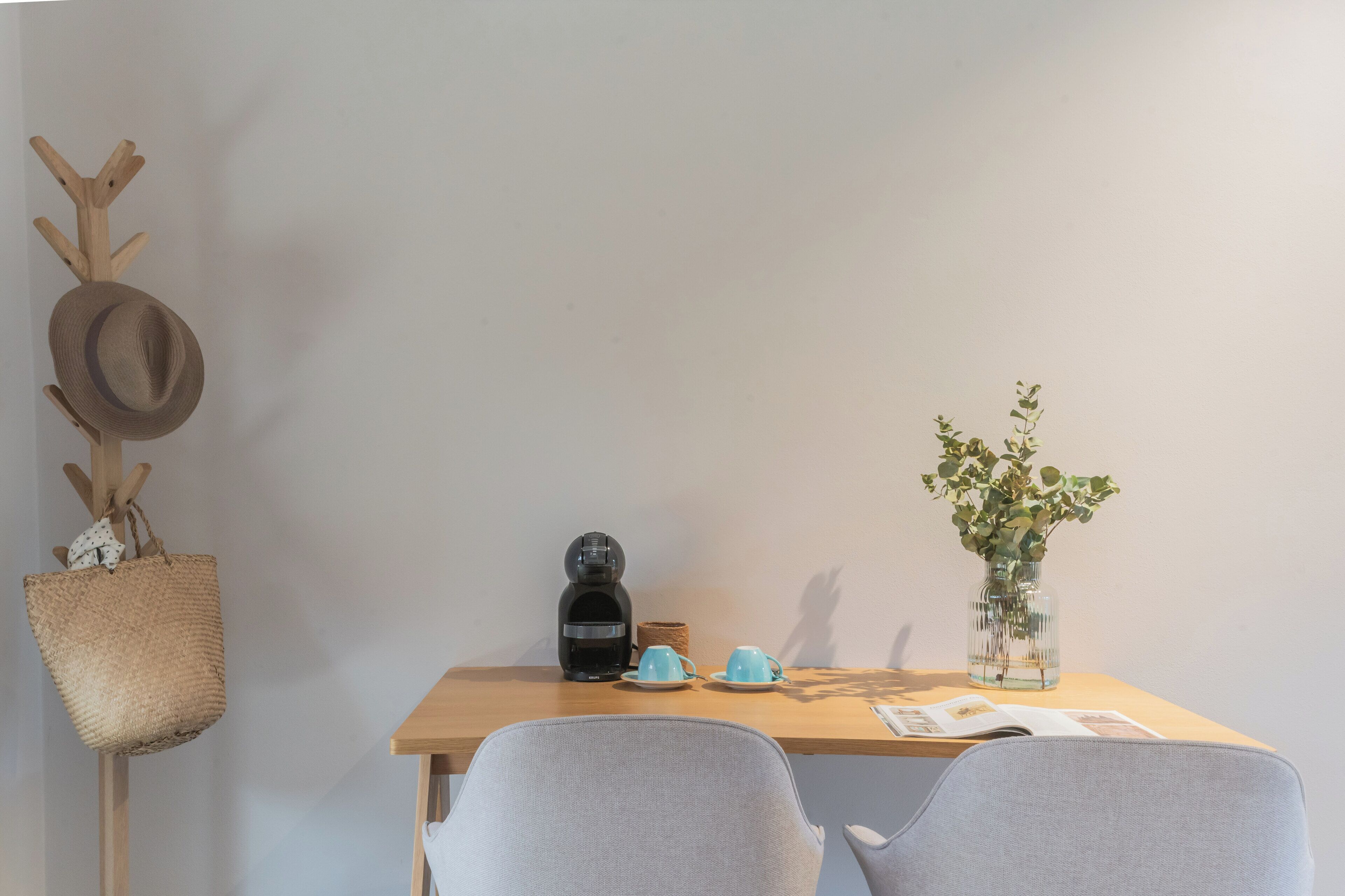 Une table en bois avec une machine à café, deux tasses bleues, un panier tressé, un vase avec des plantes vertes et un chapeau posé sur un présentoir en bois.
