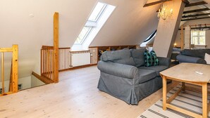 Une pièce de style loft avec un plafond en pente, des poutres en bois, un canapé gris, une table en bois et une grande lucarne.