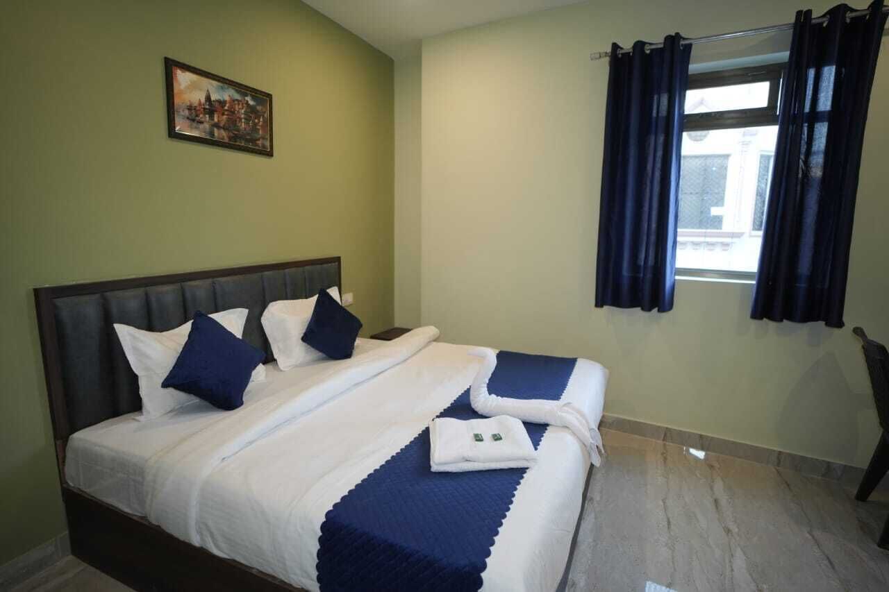 A hotel room with a double bed, two pillows, a headboard, a window with curtains, and a painting on the wall.