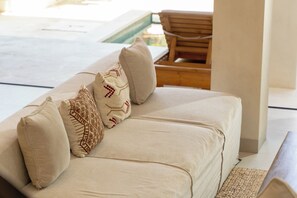 A beige sofa with patterned pillows in a bright room with a wooden chair and a view of a pool.