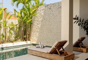Un espace aménagé au bord de la piscine, avec des chaises longues, un mur en pierre et des plantes tropicales.
