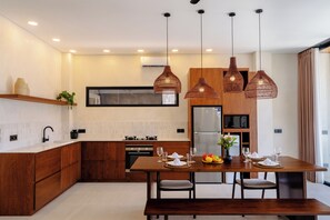 A modern kitchen with wooden cabinets, a built-in oven, and a dining area with a wooden table set for four. - Villa Balijero by Pertama Stay (Pecatu)