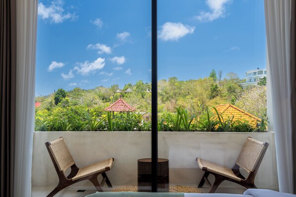 A balcony with two wicker chairs and a small round table, overlooking a lush garden and a building with a red roof. - Villa Balijero by Pertama Stay (Pecatu)