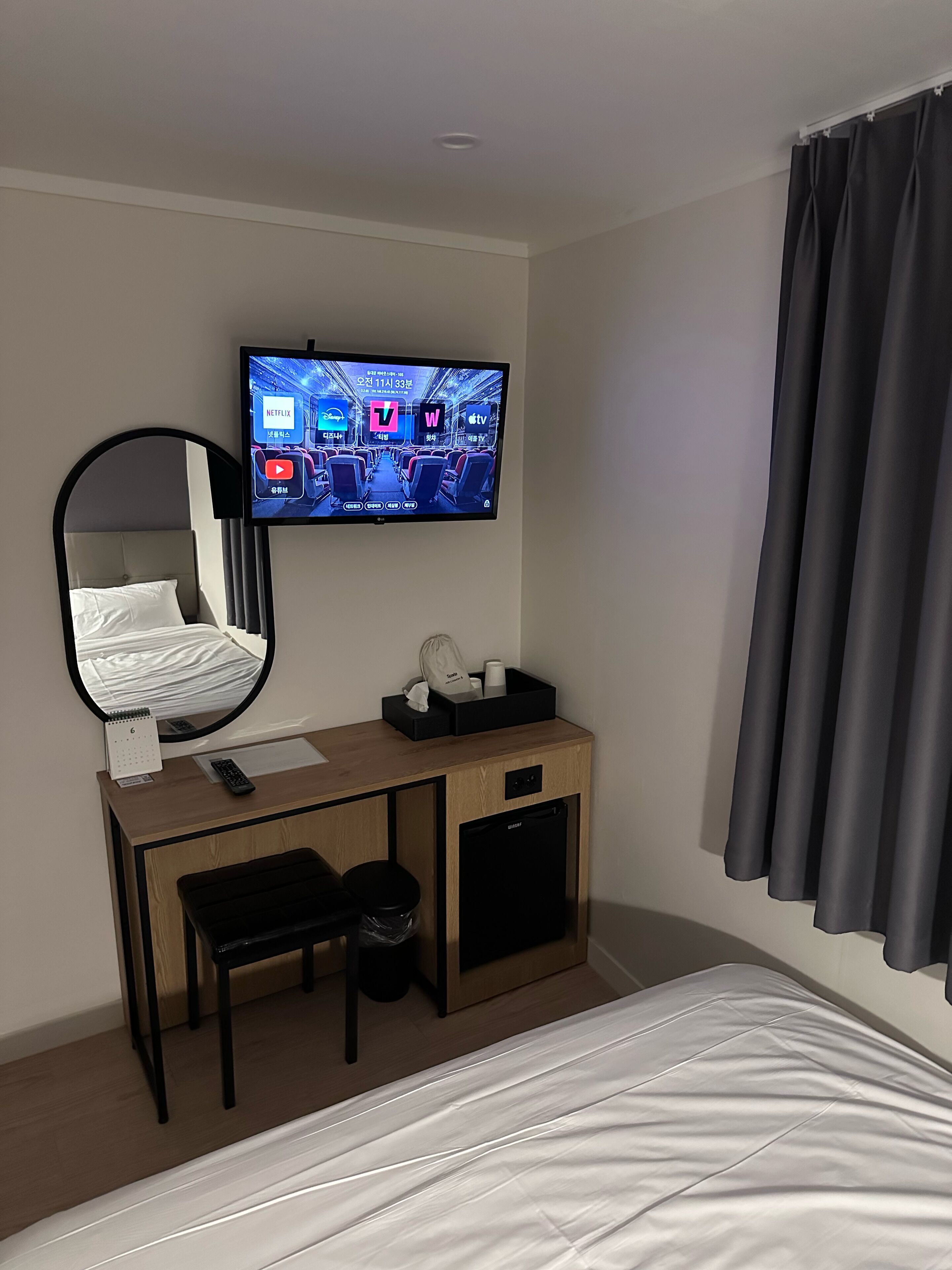 A hotel room with a bed, a desk with a mirror, a TV mounted on the wall, and a window with curtains.