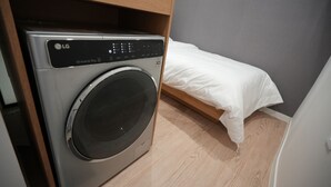 Een kleine kamer met een wasmachine, een bed en een houten plank.