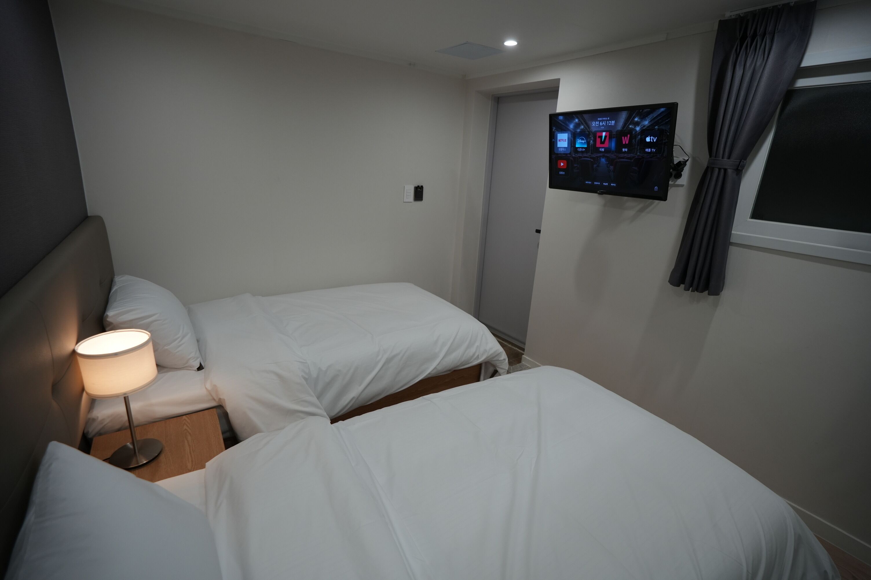 Ein Hotelzimmer mit zwei Betten, einem Nachttisch mit Lampe, einem an der Wand montierten Fernseher und einem Fenster mit Vorhängen.