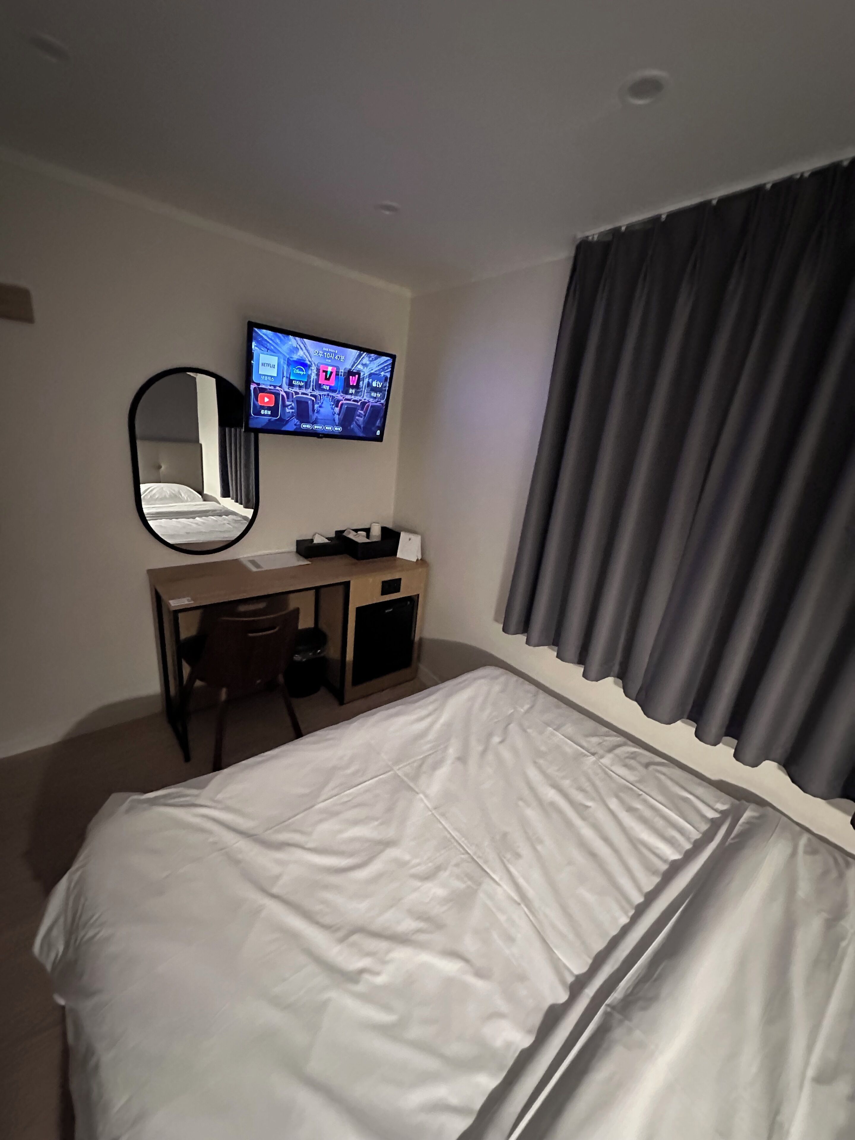 Quarto de hotel com cama, escrivaninha, cadeira, televisão fixada na parede e janela com cortinas.