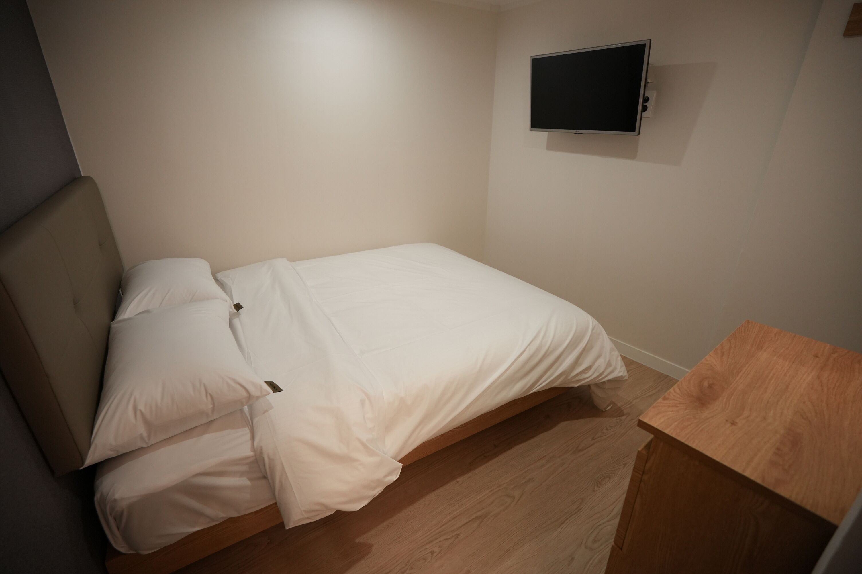 Una cama individual con ropa de cama blanca en una habitación con suelo de madera y una pequeña mesa de madera.