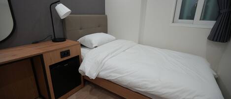 A small, modern bedroom with a single bed, a bedside table with a lamp, a built-in dishwasher, and a window with curtains.