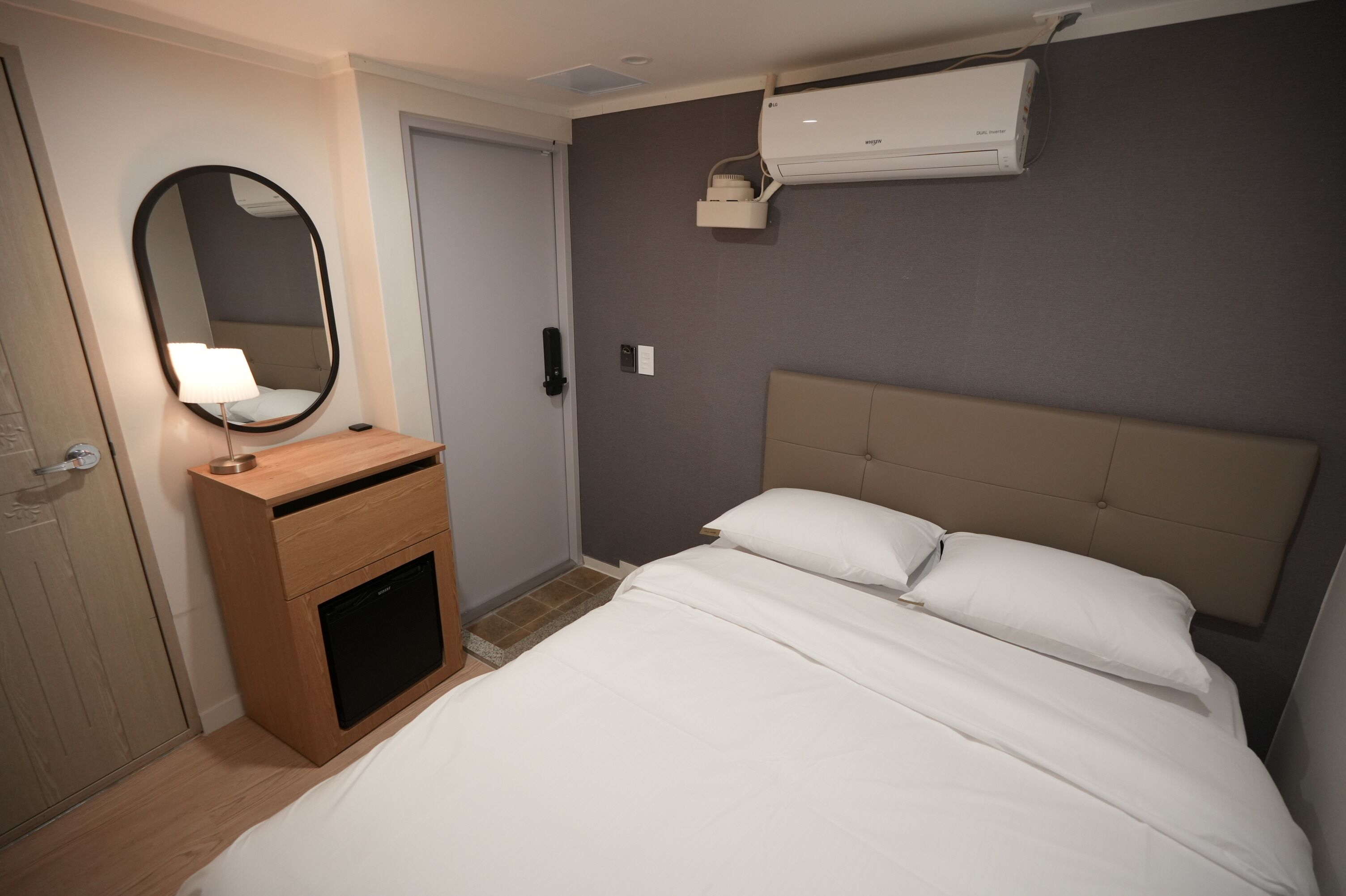 A hotel room with a bed, a bedside table with a lamp, a mirror, an air conditioning unit, and a door.
