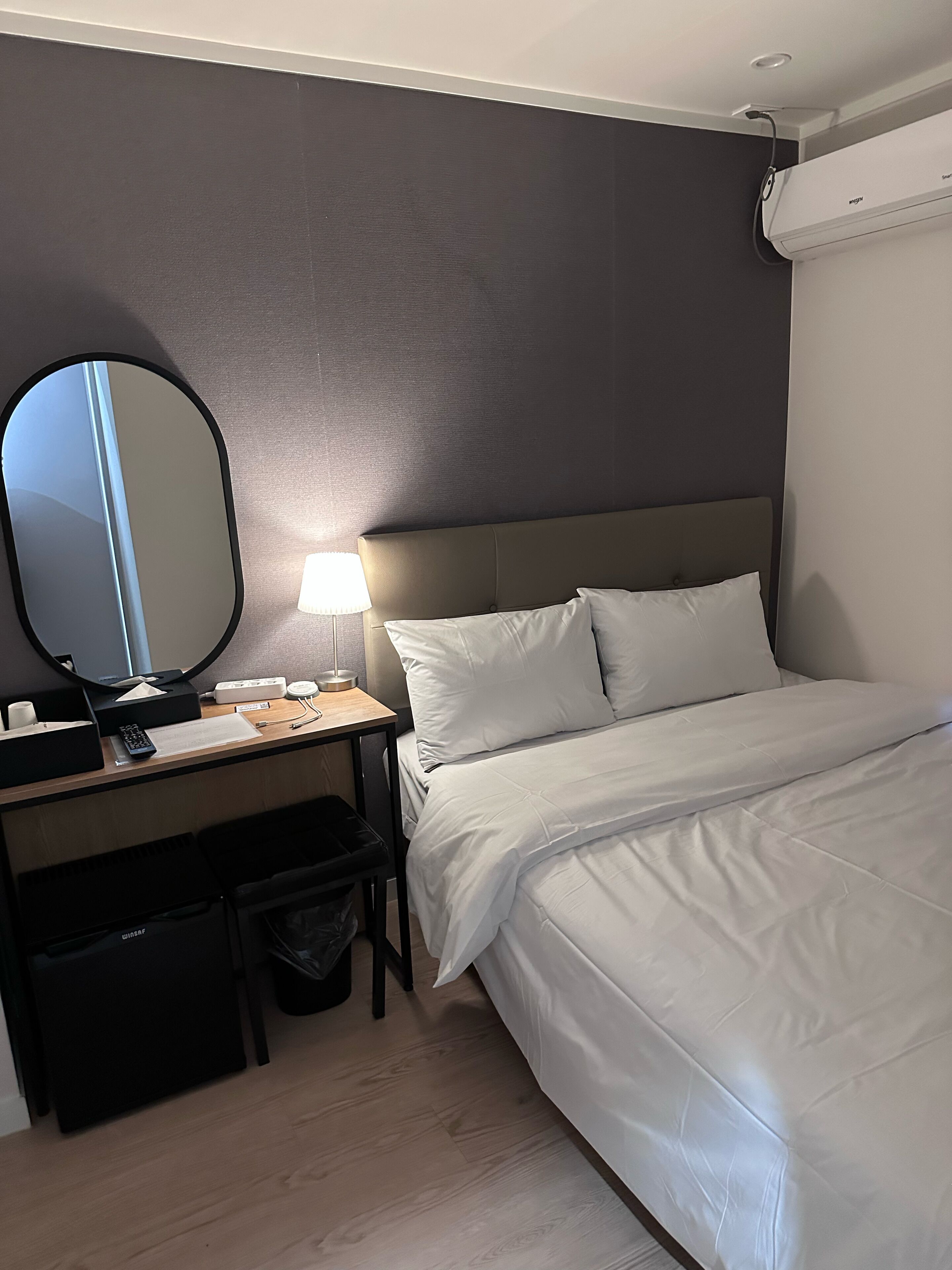A hotel room with a bed, a desk with a lamp, a mirror, and a trash bin.