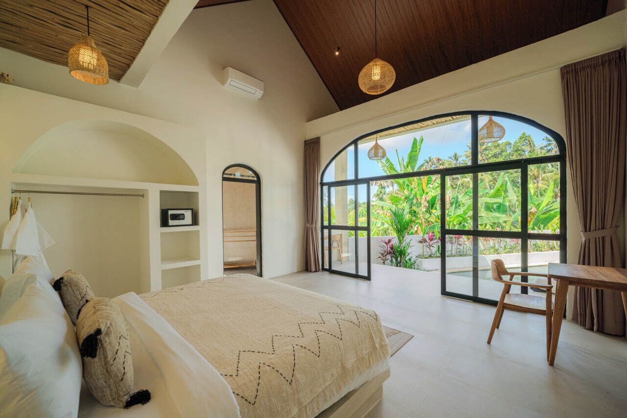 A bedroom with a large window, a bed, a wooden table, and a chair.