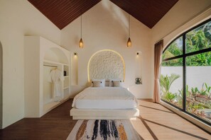 A bedroom with a large bed, a circular wall feature, pendant lights, a view of greenery, and a wooden ceiling. - Casa Mahesa Villas (Ubud)