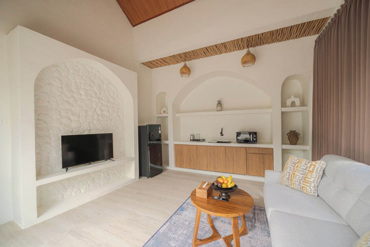 Un salon moderne avec un téléviseur à écran plat fixé sur un mur texturé, une table basse en bois sur laquelle sont disposés des fruits, un canapé agrémenté de coussins à motifs et un coin cuisine avec des meubles en bois et un four à micro-ondes.