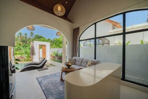 A modern living room with a curved sofa, a coffee table, and a view through a large window. - Casa Mahesa Villas (Ubud)