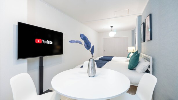 Una camera d'albergo moderna con televisione a schermo piatto montata a parete, un tavolo rotondo bianco con un vaso, sedie bianche e un letto con cuscini blu e verdi.