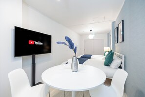 A modern hotel room with a flat-screen TV mounted on the wall, a round white table with a vase, white chairs, and a bed with blue and green pillows. - TRINITY GOLD (Osaka)