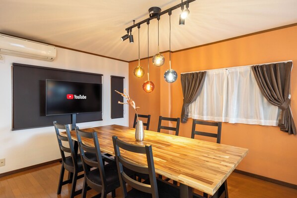 A conference room with a wooden table, black chairs, a flat-screen TV, pendant lights, and a window with curtains.