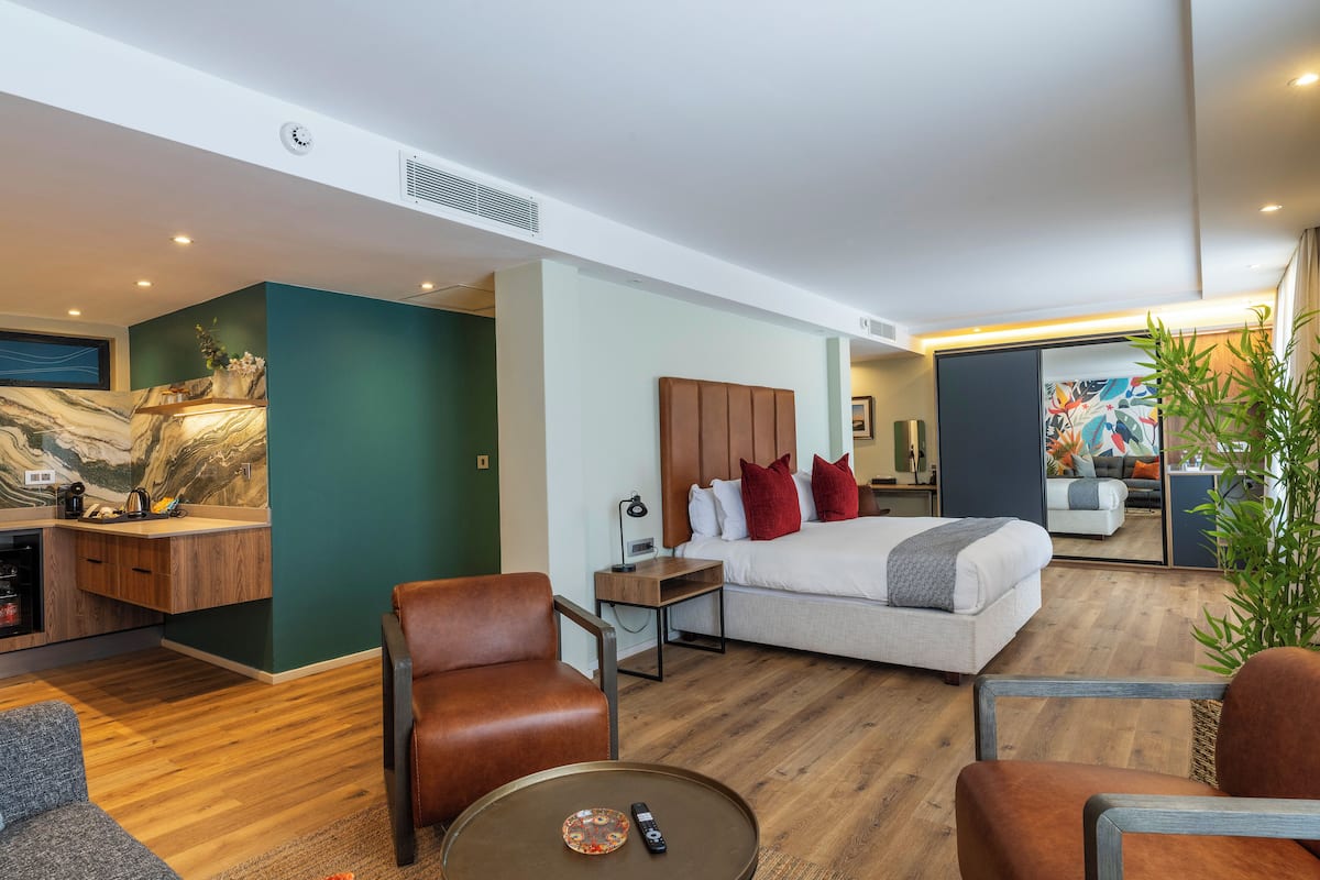 A modern hotel room with a bed, two chairs, a small table, a desk with a lamp, a kitchen area, and a large mirror.
