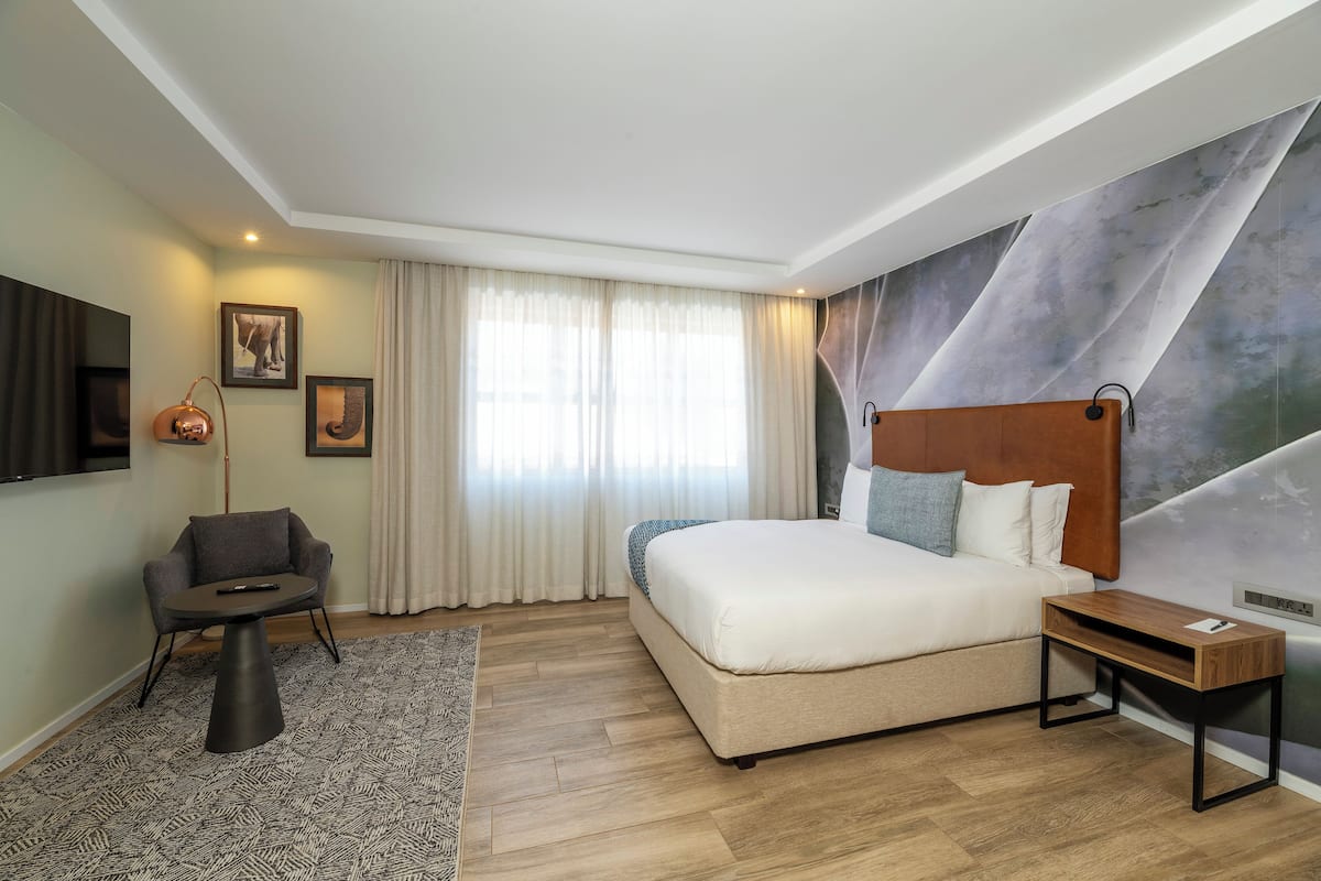 A modern hotel room with a large bed, a small desk, a chair, a TV, and a wall mural.